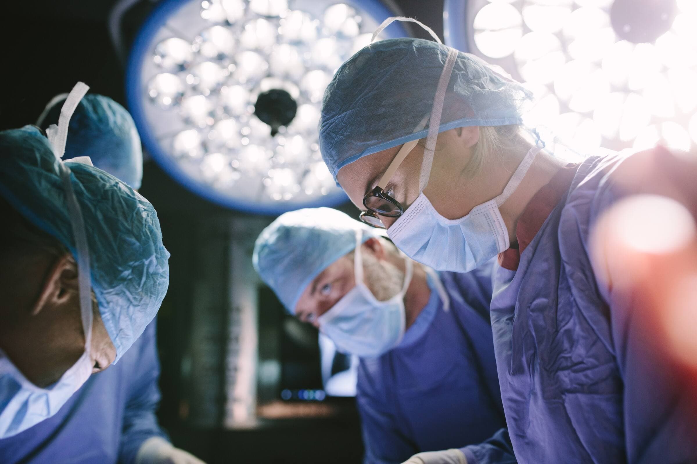 A surgeon in a cap and mask at work under operating lights